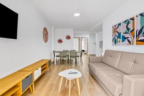A modern living room with a sofa, a wooden TV stand, a dining area with chairs, and a small white table.