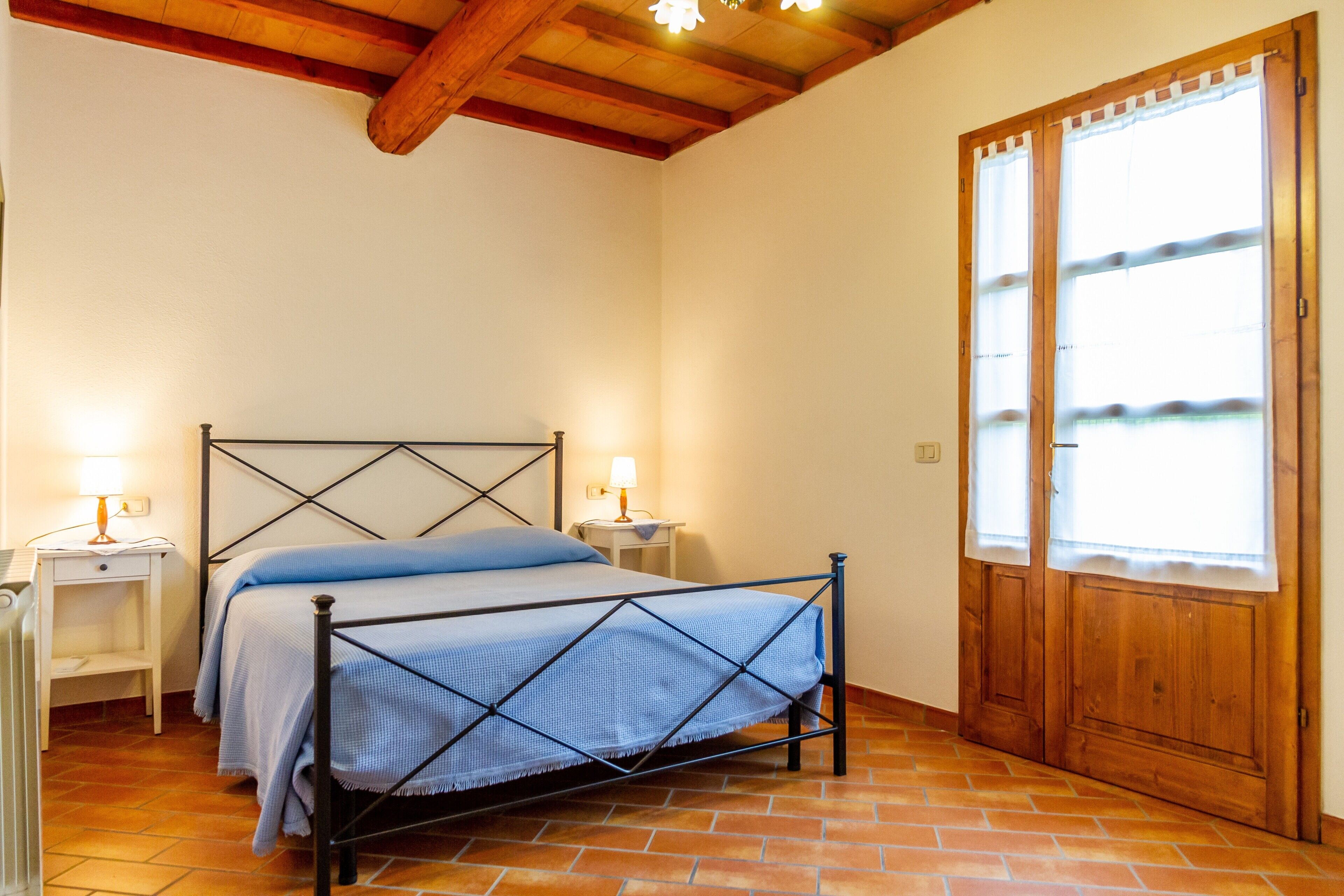 A bedroom with a metal bed frame, two bedside tables with lamps, a wooden door, and a tiled floor.
