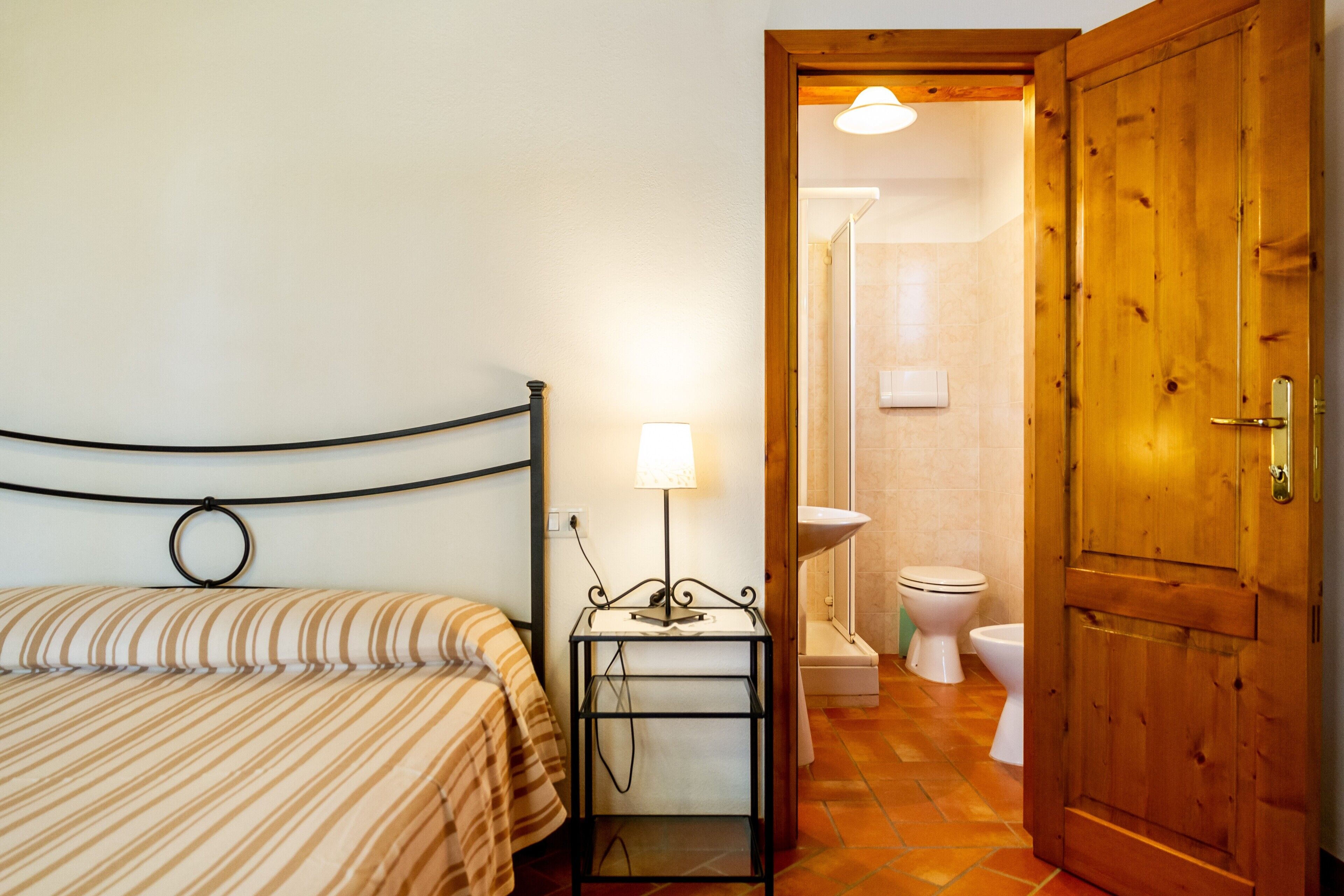 A bedroom with a bed, a bedside table with a lamp, a wooden door, and a bathroom with a toilet and sink.