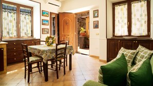 Zona pranzo con tavolo apparecchiato per quattro, sedie in legno, un divano verde e angolo cottura con pensili e finestra con tende.