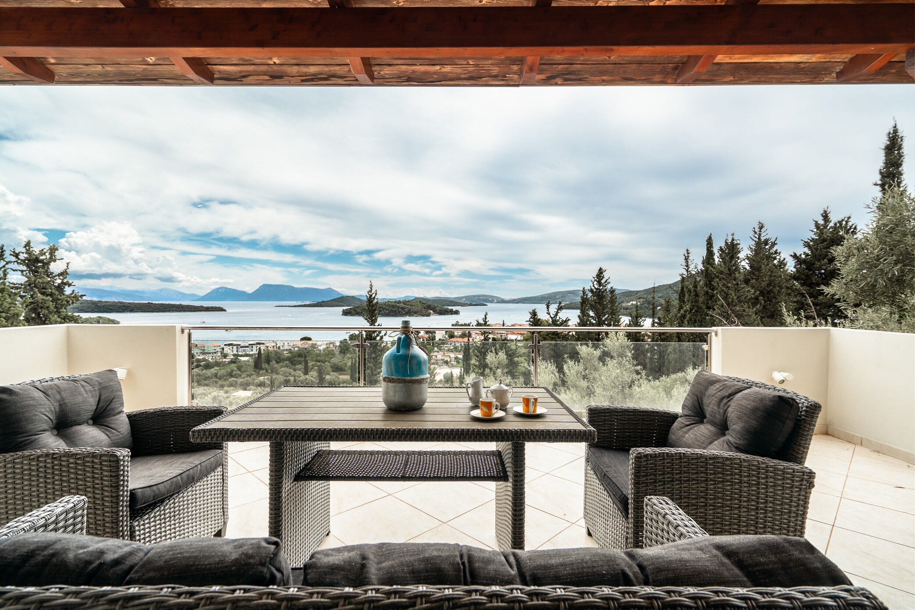 Uma varanda com móveis de vime, uma mesa com um vaso e vista para um lago e montanhas.