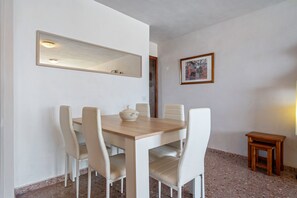 Un coin repas avec une table en bois et des chaises blanches, un petit tabouret en bois, un tableau encadré au mur et un miroir au-dessus de la table.