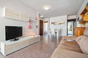 Una sala de estar moderna con un televisor de pantalla plana, un mueble blanco para entretenimiento, un sofá color beige y un comedor con mesa y sillas blancas.
