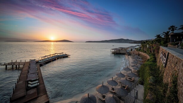 A coastal resort with a wooden pier extending into the sea, beach umbrellas, and a stone wall with a sign. - Swissôtel Living Bodrum (Bodrum)