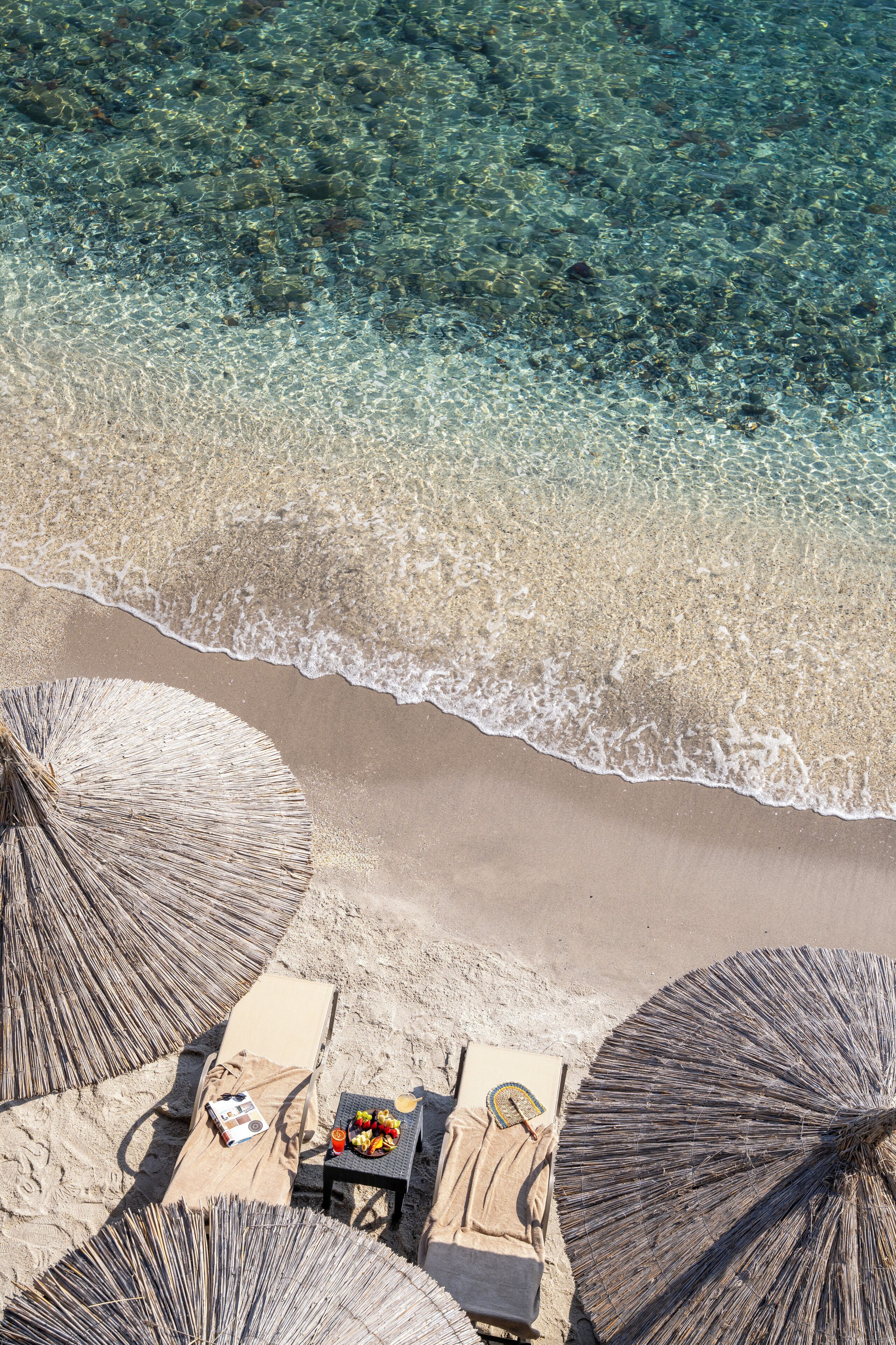 Vista aérea de una playa con agua cristalina, una orilla arenosa y un área de descanso con una mesa, sillas y sombrillas.