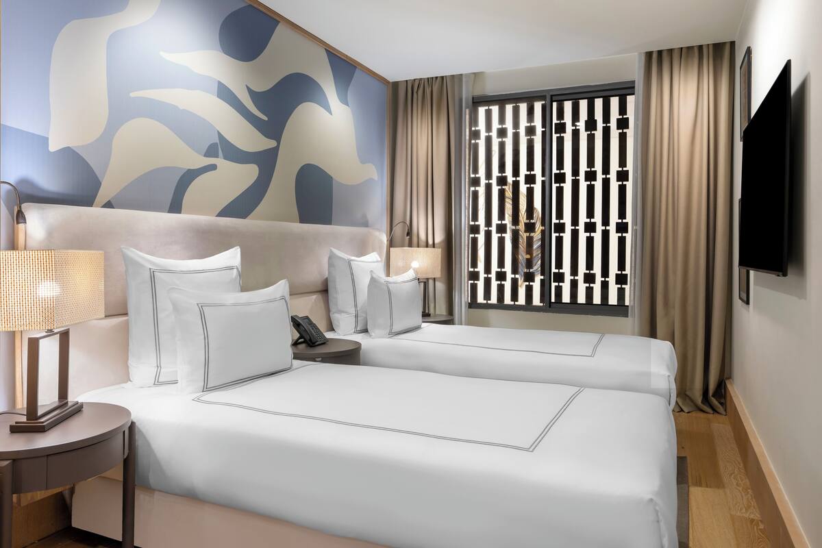 A hotel room with two beds, a large abstract wall mural, a TV, and a window with a decorative screen.