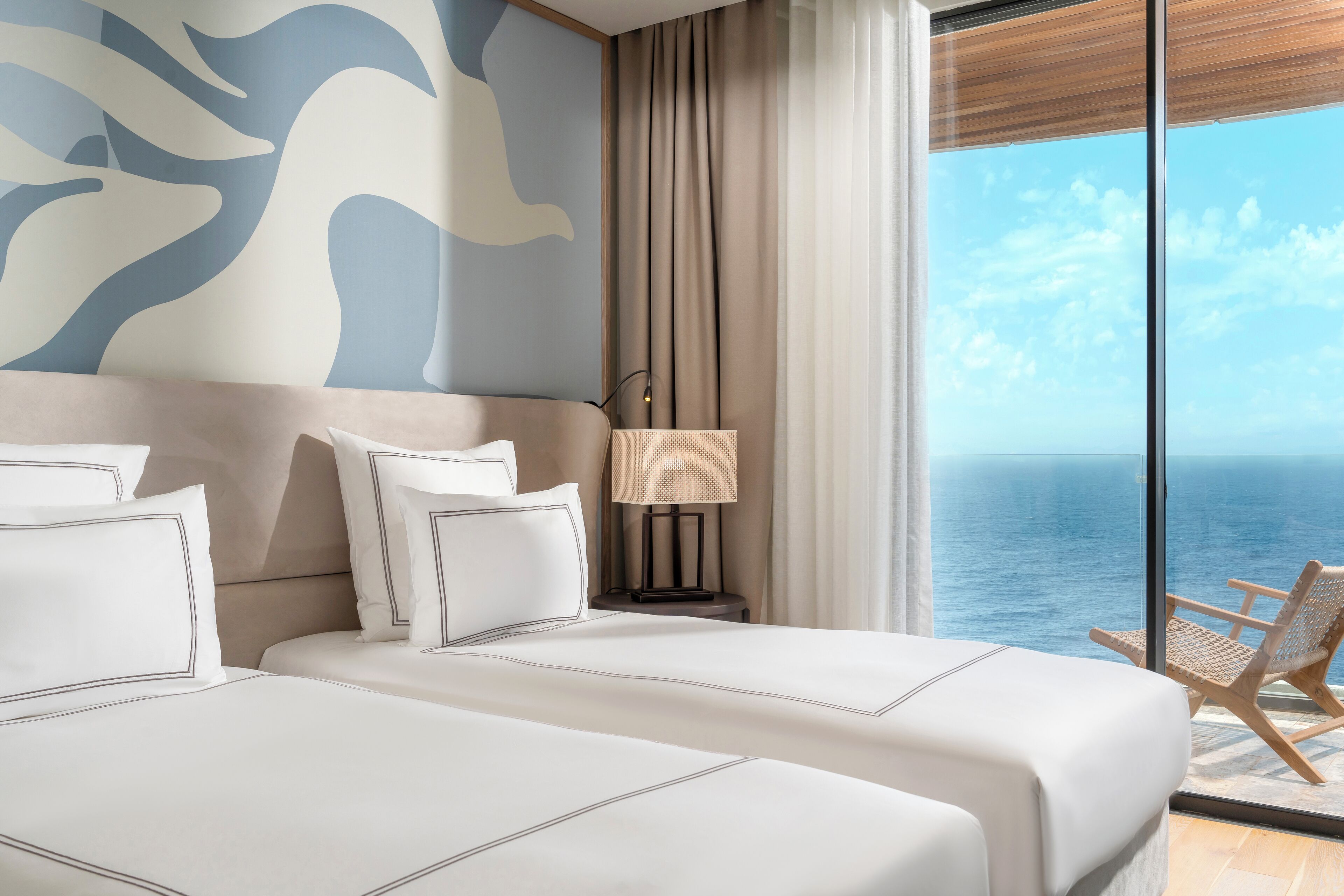 A bedroom with two beds, a nightstand with a lamp, a view of the sea, and a chair by the window.