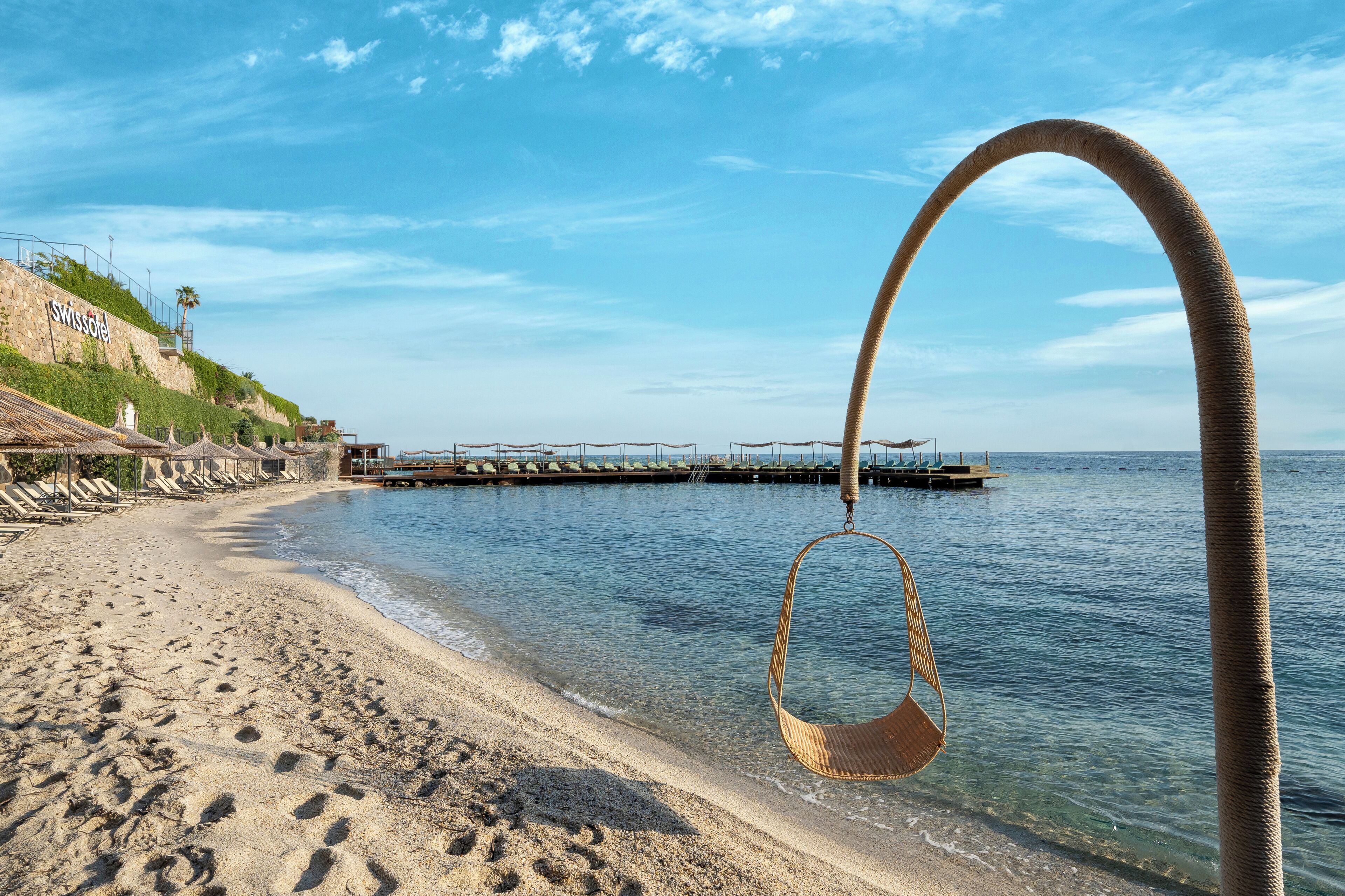 A beach with a swing chair hanging from a curved structure, a pier extending into the water, and a building with a sign that reads "Swissotel".