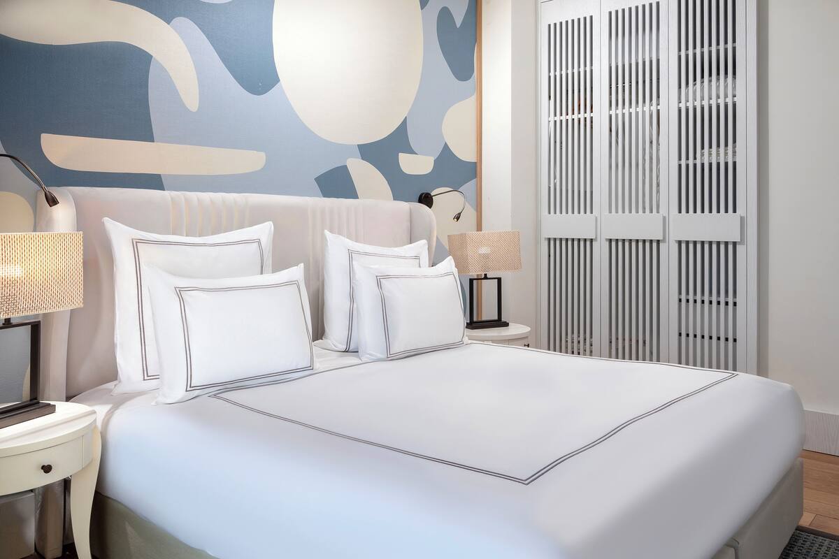 A neatly made bed with white linens and a modern headboard, a bedside lamp, and a wall with abstract art.