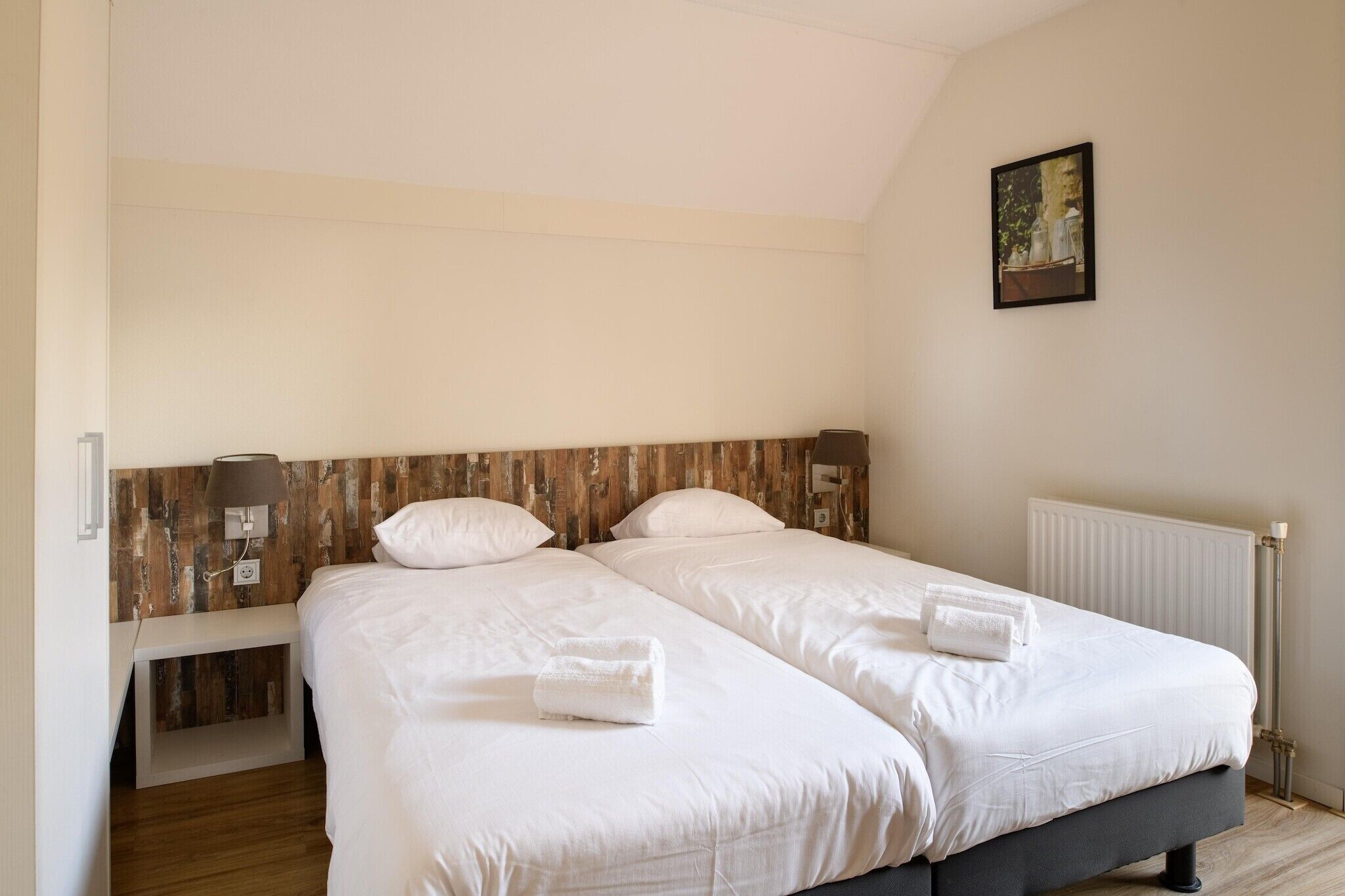 Une chambre double avec deux lits, une tête de lit en bois, une table de chevet, une lampe et un tableau encadré au mur.