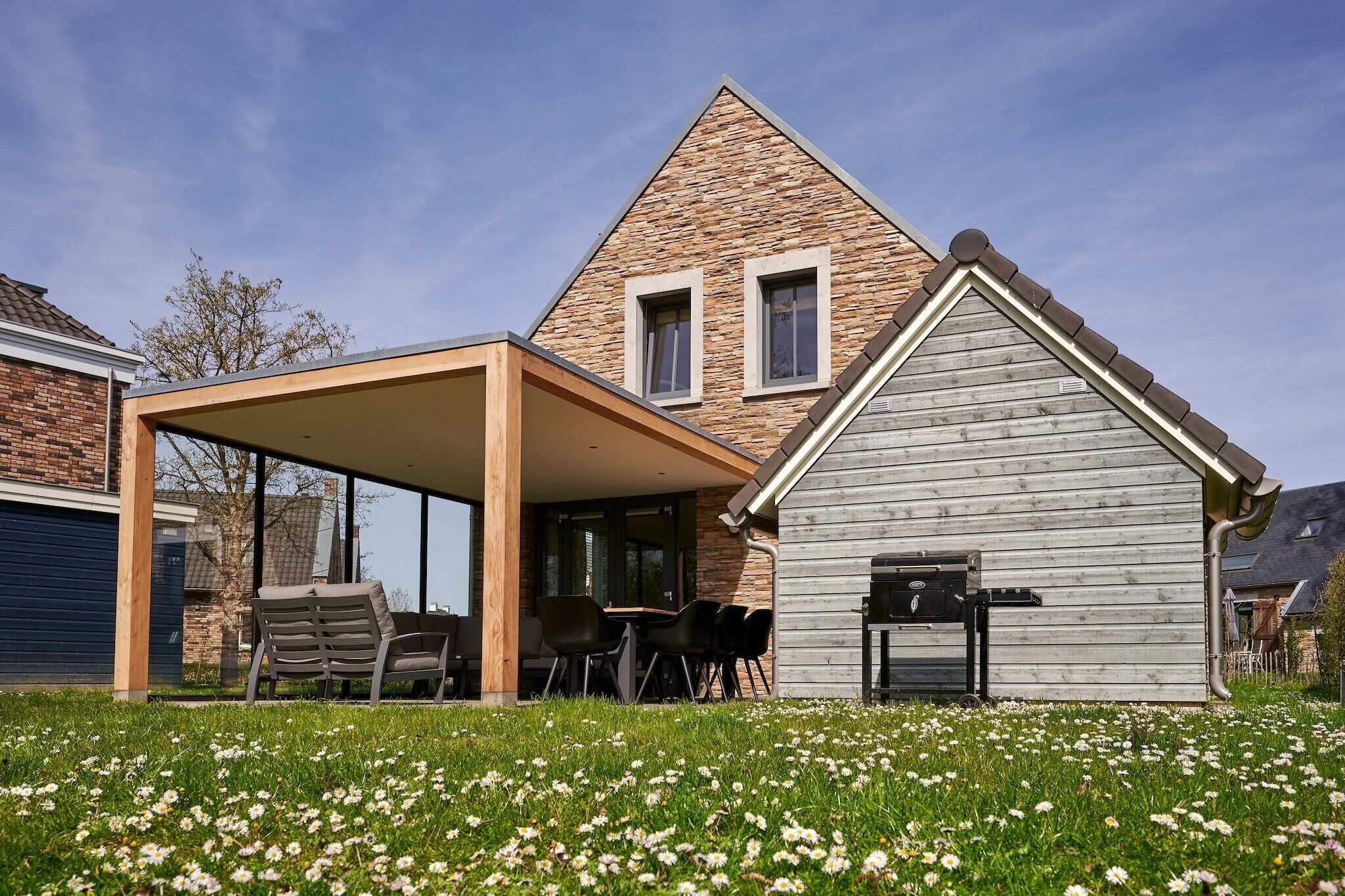 Een modern huis met een gevel van natuursteen, een overdekt terras met tuinmeubilair en een kleine schuur met een barbecue.