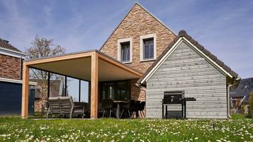 Een modern huis met een gevel van natuursteen, een overdekt terras met tuinmeubilair en een kleine schuur met een barbecue.