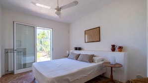 Un dormitorio con una cama, ventilador de techo, una mesita pequeña y vista a palmeras a través de un ventanal.