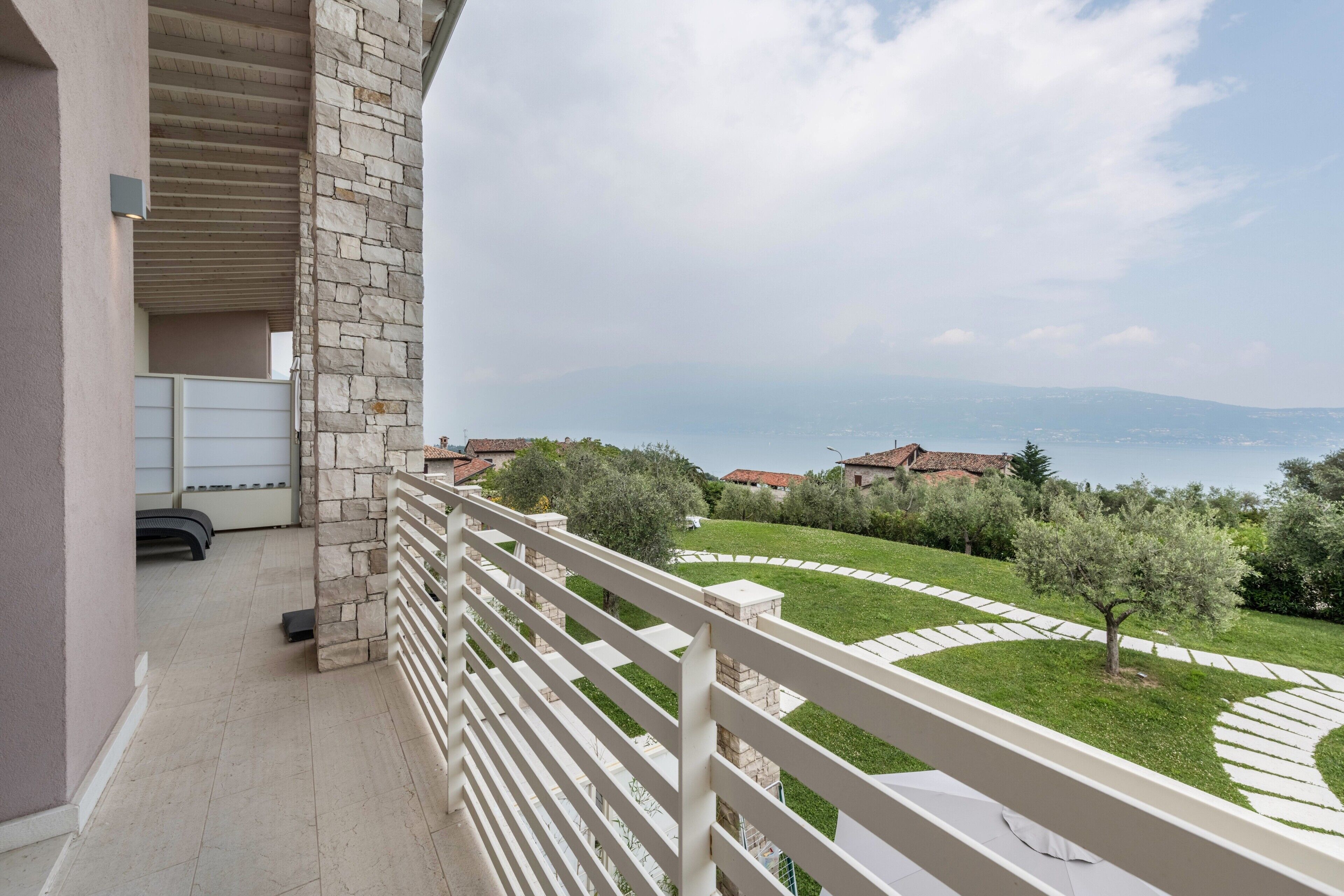Un balcon doté d’un mur en pierre, d’une balustrade blanche et offrant une vue sur un jardin aménagé, avec des arbres et une chaîne de montagnes au loin.