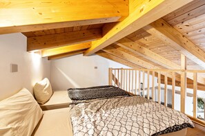Ein Schlafzimmer mit schrägen Wänden, Holzbalken, einem Bett mit gemusterten Betttextilien und Blick ins Freie.