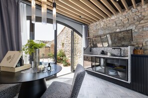 An outdoor kitchen with a stone wall backdrop, a round table with a vase and candles, and a seating area with patterned cushions.