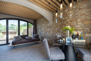 A modern interior with a stone wall, a wooden ceiling, a sofa, patterned chairs, and a table with wine bottles.