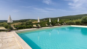 En swimmingpool med solstolar och parasoller, omgiven av grönska och kullar.
