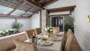 An outdoor dining area with a stone wall, wooden beams, wicker chairs, a table set with plates, glasses, and candles. - Villa 'Casa Al Mare' with Private Terrace, Wi-Fi and Air Conditioning (Siracusa)