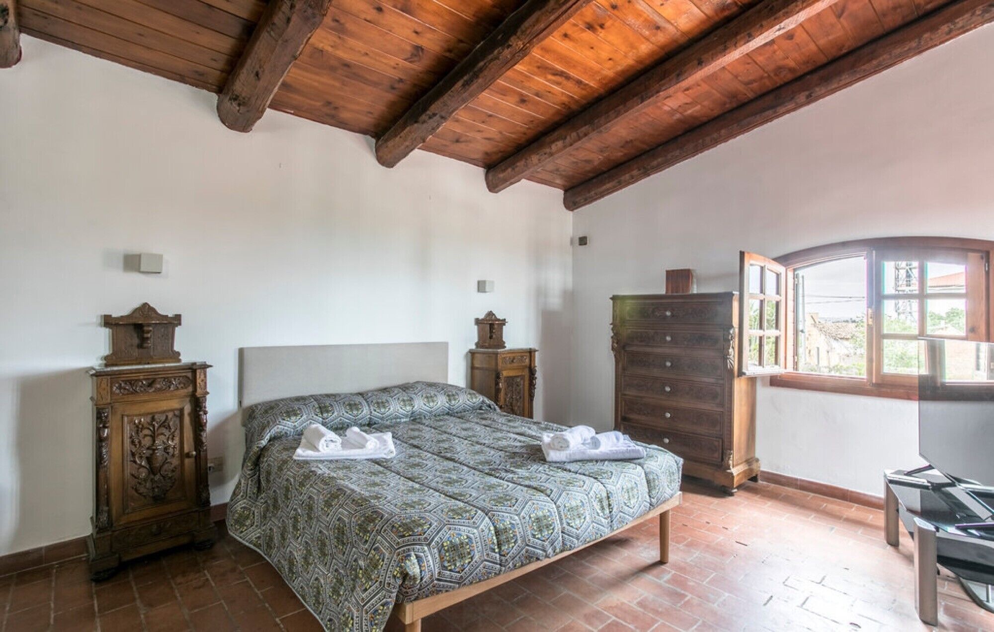 Une chambre avec un plafond en bois, un lit avec une literie à motifs, une commode et une fenêtre.