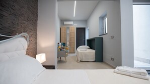 A modern hotel room with a large bed, a desk, a chair, and a small table. There is a window providing natural light and a view of the outside. - Description (Catania)
