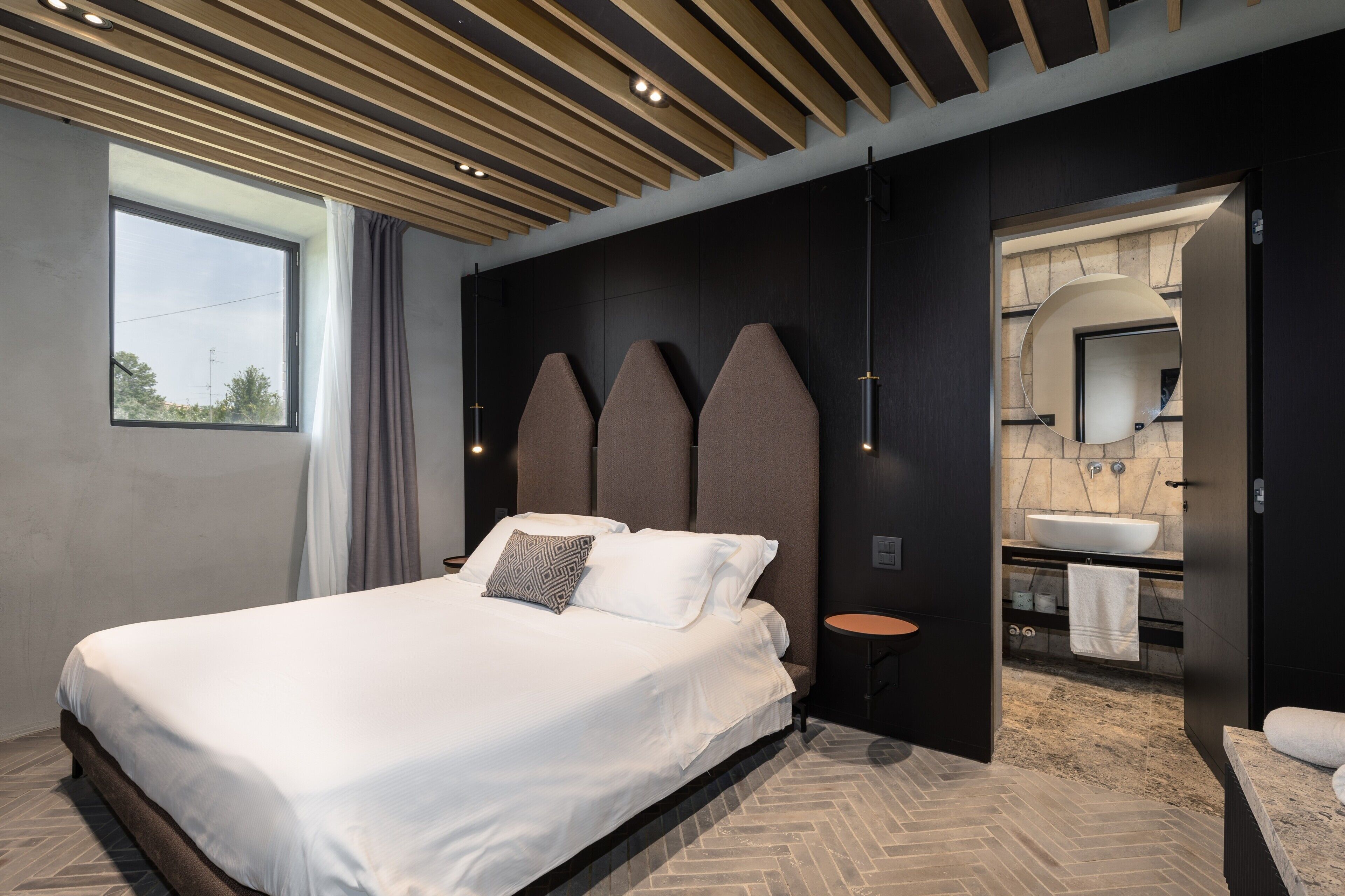 Een moderne hotelkamer met een groot bed, een klein rond tafeltje, een inbouwsysteem voor de wastafel en een spiegel in de badkamer.