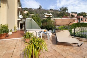 Une terrasse agrémentée de plantes en pot, une table avec des chaises, un transat et une vue sur les bâtiments environnants.