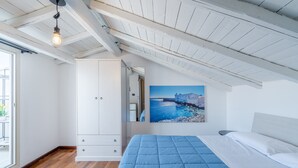 A bedroom with a wooden floor, a bed with a blue quilt, a white wardrobe, and a painting of a coastal scene. - Holiday Apartment 'Cheyenne's House' with Terrace, Balcony & Wi-Fi (Siracusa)