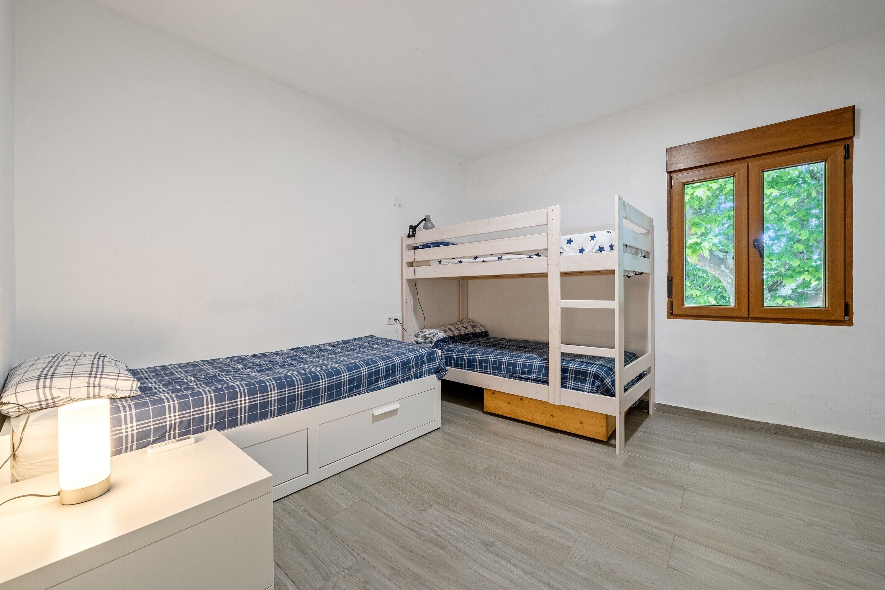 A bedroom with bunk beds, a bedside table with a lamp, and a window with a view of greenery.