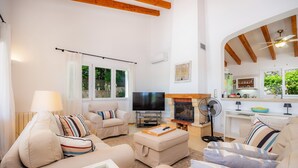 A living room with a TV, sofa, ottoman, fireplace, and kitchen area. - Villa 'Victoria' with Private Pool, Wi-Fi and Air Conditioning (Crestatx)