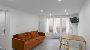 Ein modernes Wohnzimmer mit einer Couch, einem Esstisch, Stühlen, einem an der Wand montierten Fernseher und einer Glastür, die zu einem Außenbereich führt.