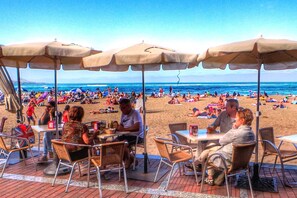 Un café situé en bord de mer, avec des tables à l’extérieur et des parasols, offrant une vue sur une plage très fréquentée.