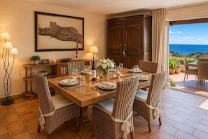 Un coin repas avec une table en bois dressée pour six personnes, des chaises en osier, un grand tableau encadré représentant un léopard et une vue sur l’océan par une porte coulissante.