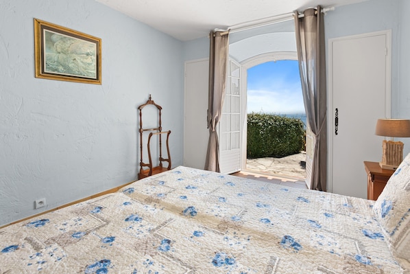 Ein Schlafzimmer mit Bett, einem Gemälde, einem Sessel und Blick ins Freie.