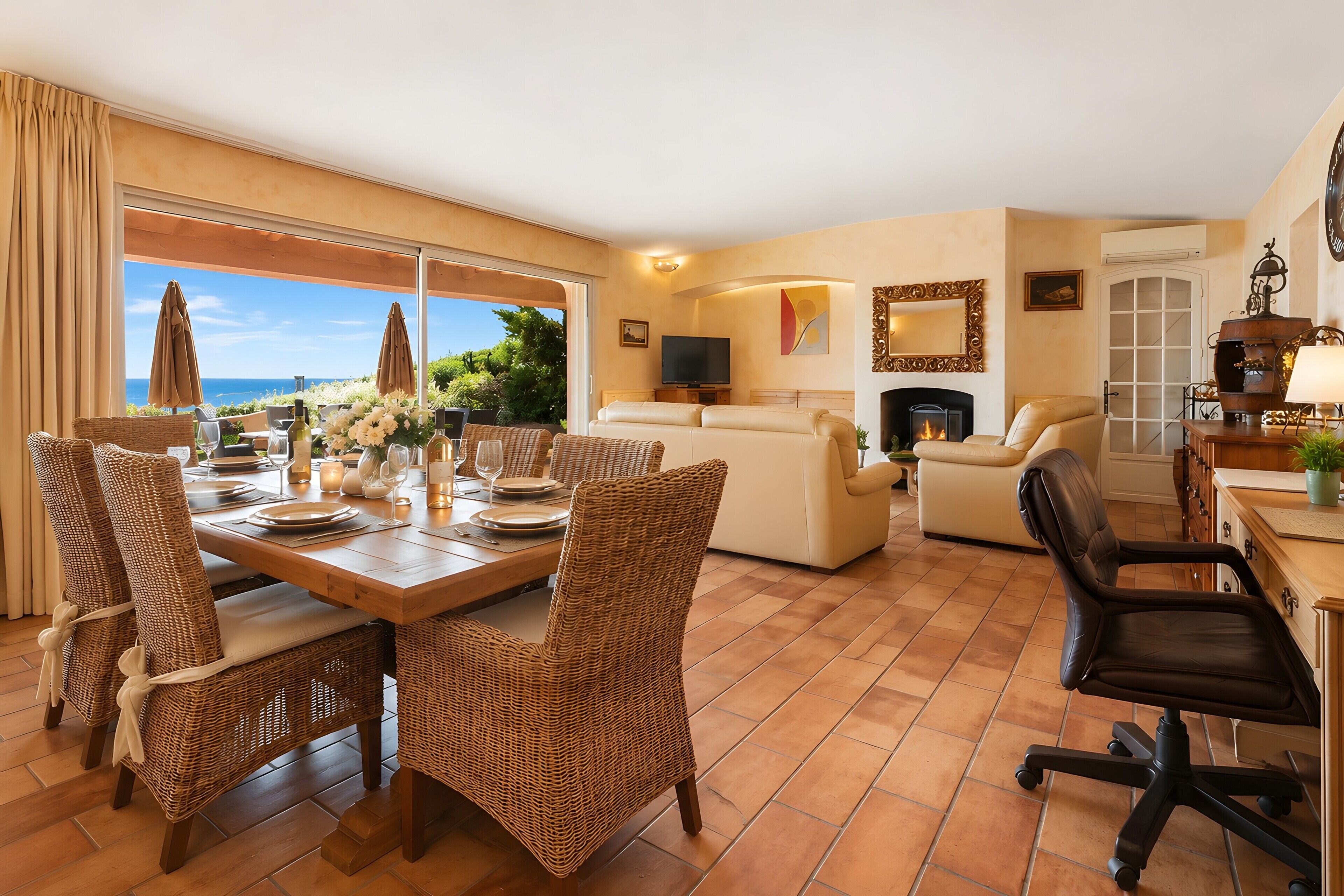 Een ruime woonkamer met een eettafel voor vier personen, omringd door rieten stoelen. Achtergrond bevat een bank en een open haard.