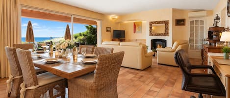 A spacious living area with a dining table set for four, surrounded by wicker chairs. A sofa and a fireplace are visible in the background.