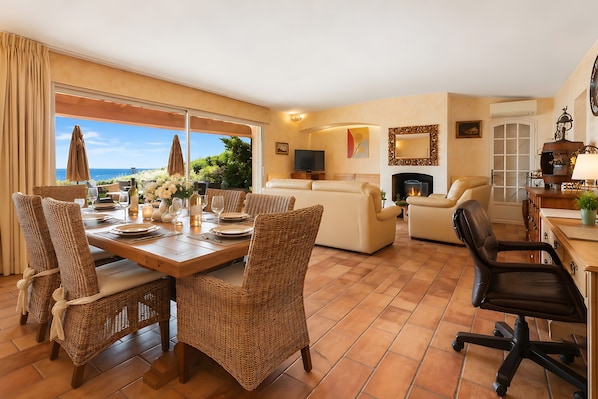Un vaste espace de vie comprenant une table à manger pour quatre personnes, entourée de chaises en osier. On aperçoit un canapé et une cheminée au fond de la pièce.