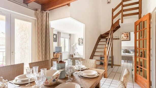 Comedor con una mesa de madera preparada para una comida, una escalera y vistas al espacio de estar contiguo.