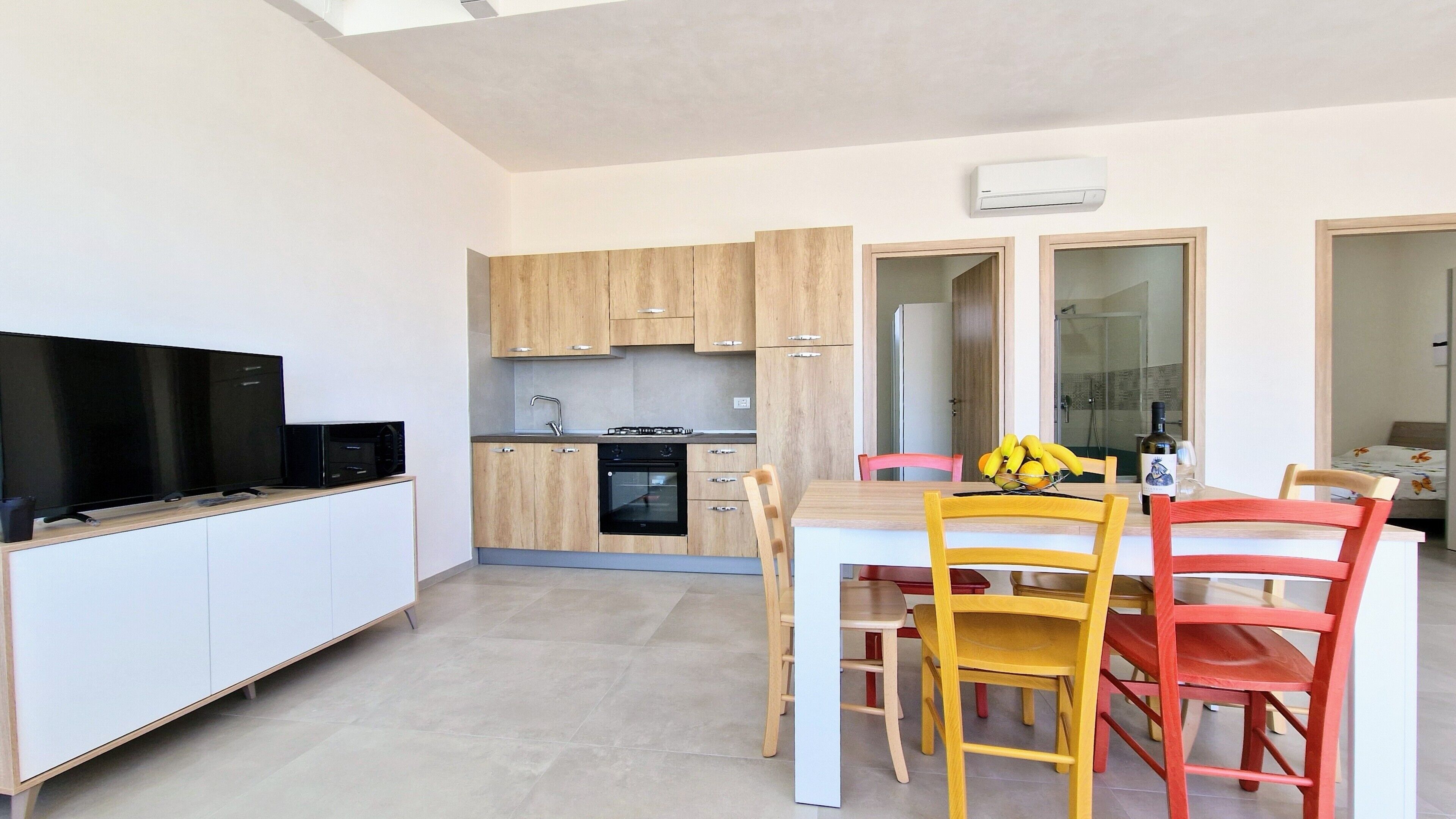Cocina moderna con armarios de madera, comedor con mesa y sillas, y televisor sobre un mueble.