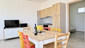 A modern kitchen with wooden cabinets, a dining table with chairs, a TV, and a bed in the adjacent room.