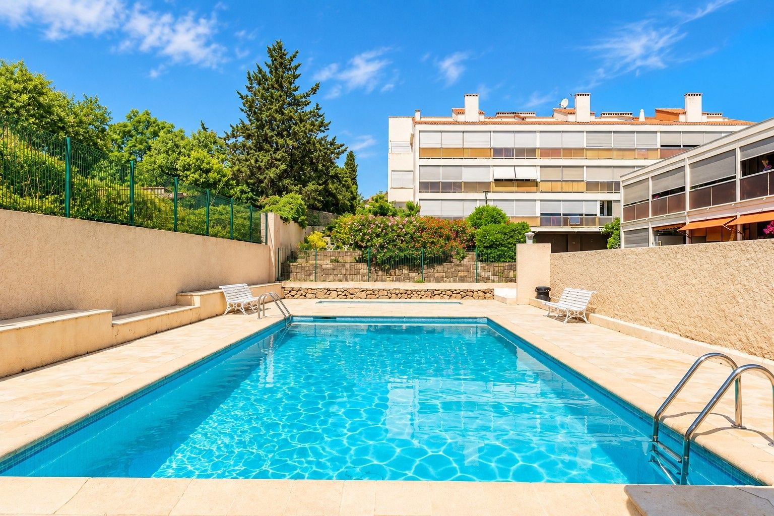 Une piscine rectangulaire avec des marches permettant d’y accéder, entourée d’un petit muret en pierre et d’une clôture en métal.