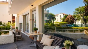 Un área exterior con mesas, sillas y un banco, con vistas a una calle con edificios y zonas verdes.