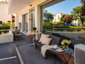 An outdoor seating area with tables, chairs, and a bench, overlooking a street with buildings and greenery.