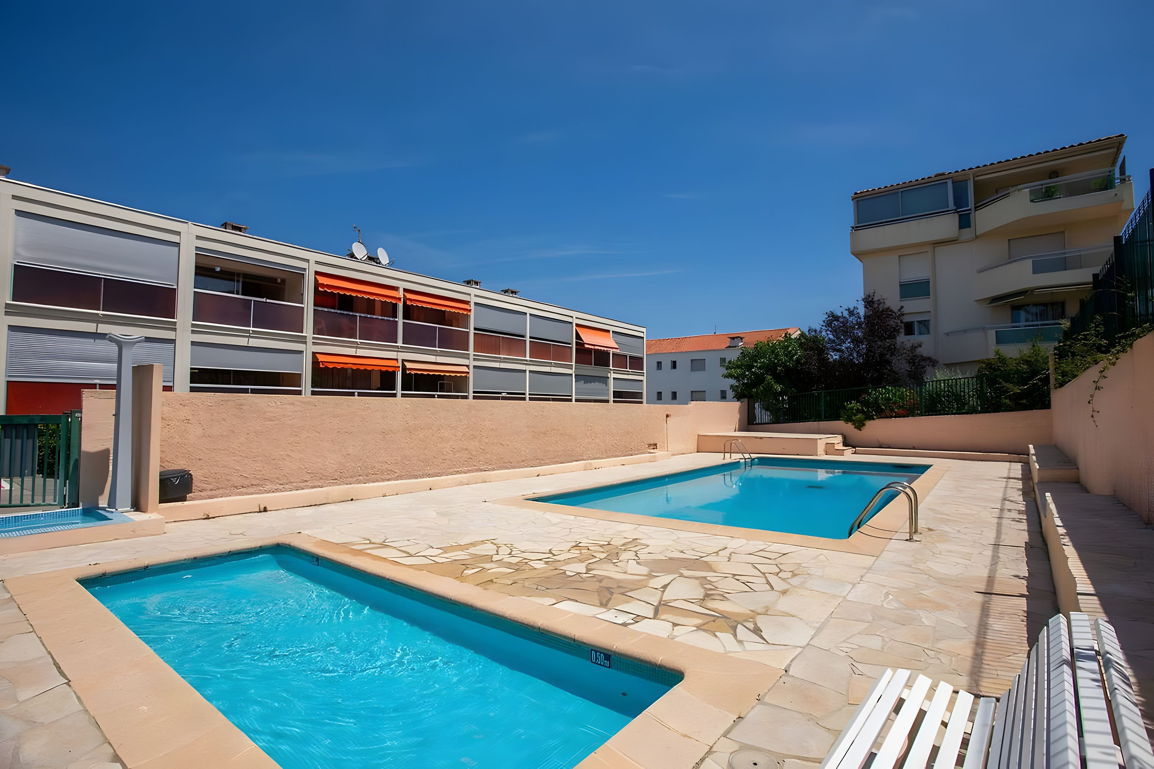 Un espace piscine comprenant deux piscines, un espace détente couvert et un bâtiment doté de balcons.