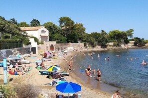 Una playa con gente nadando y tomando el sol, sombrillas y un edificio con un muro de piedra.