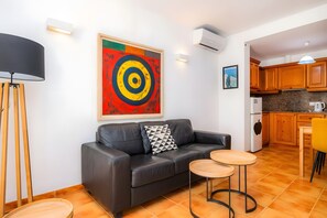 A modern living room with a black leather sofa, a colorful abstract painting, a small wooden coffee table, and a kitchen area with wooden cabinets and a tiled floor.