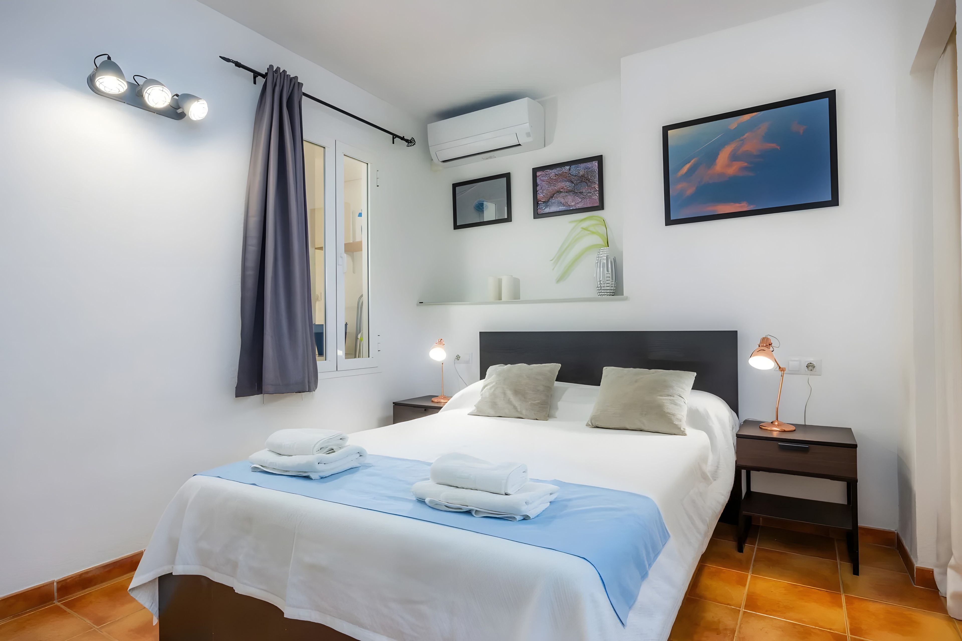 A hotel room with a bed, bedside lamps, a nightstand, a wall-mounted air conditioner, and a framed picture.