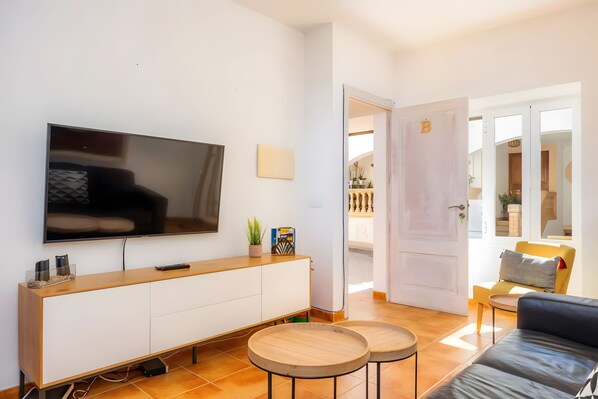 Una sala moderna con un televisor de pantalla plana, un mueble de madera, dos mesas redondas, un sofá y vistas al balcón.