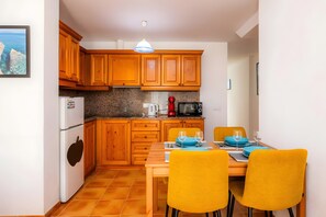 A compact kitchen with wooden cabinets, a dining table with yellow chairs, and a small island with a kettle and coffee maker.