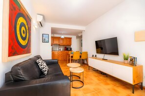 A modern living room with a black leather sofa, a dining area with yellow chairs, a kitchen with wooden cabinets, and a mounted flat-screen TV.